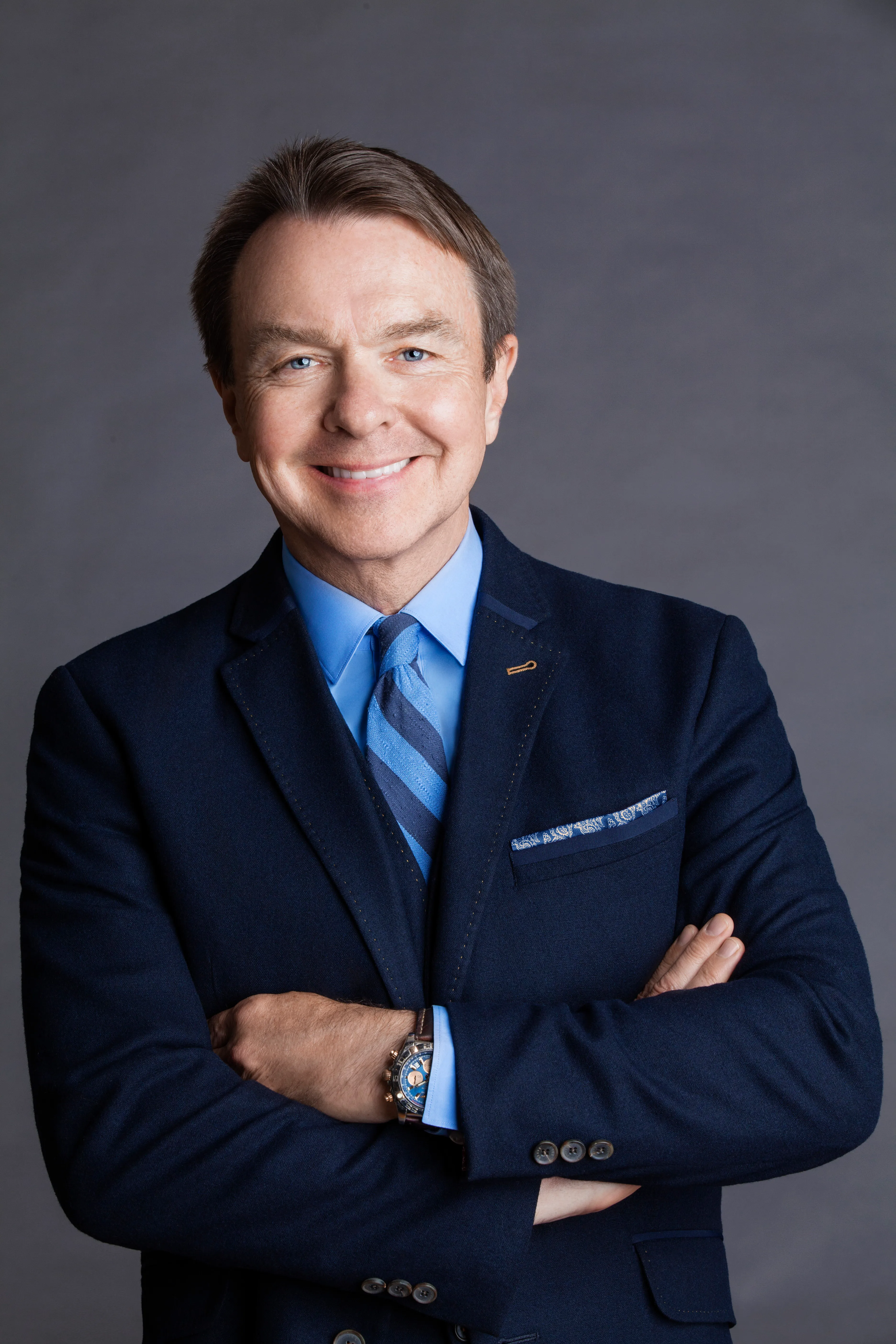 Smiling middle-aged man with blue eyes wearing a navy blazer, striped blue tie, and blue shirt with arms crossed.